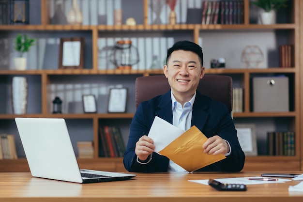 Portret van een succesvolle Aziatische zakenman ontving een goedkeuringsbrief van de bank om de kredietlimiet te verlengen een gelukkige man aan het werk aan de tafel met een envelop en kijkend naar de camera