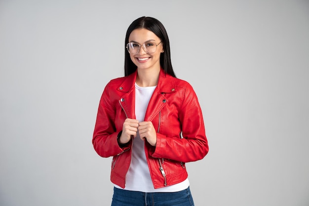 Portret van een succesvolle jonge vrouw in een bril die staat en naar de camera kijkt met een brede glimlach. Indoor studio-opname, geïsoleerd op een grijze achtergrond