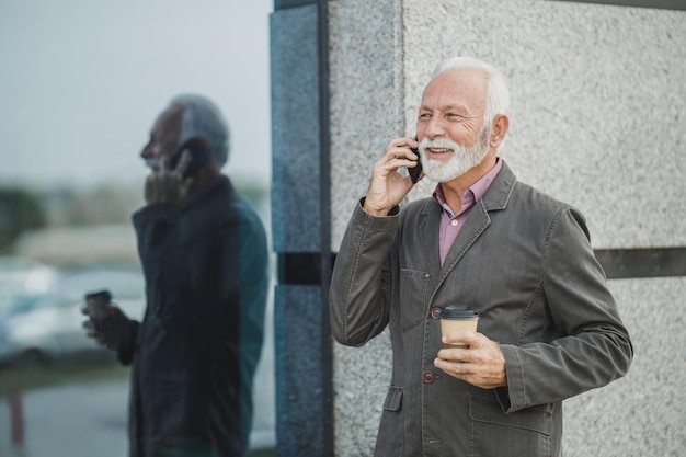 Portret van een succesvolle senior zakenman die op een smartphone praat terwijl hij een koffiepauze heeft voor een bedrijfsgebouw.