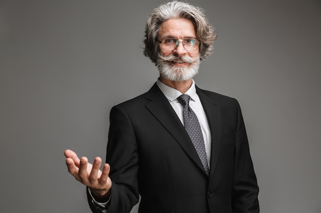 Portret van een succesvolle volwassen zakenman met een formeel zwart pak en een bril die naar de camera glimlacht met gebarende hand geïsoleerd over grijze muur