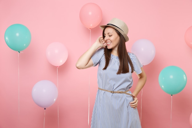 Portret van een tedere fascinerende jonge vrouw met een stro-zomerhoed en een blauwe jurk die de hand in de buurt van het gezicht houdt op een roze achtergrond met kleurrijke luchtballonnen. Verjaardag vakantie feest mensen oprechte emoties.