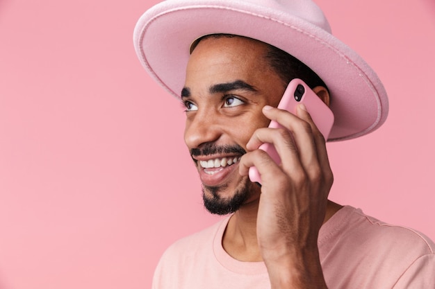 Portret van een tevreden Afro-Amerikaanse man met een hoed die op een mobiele telefoon praat en glimlacht, geïsoleerd op roze