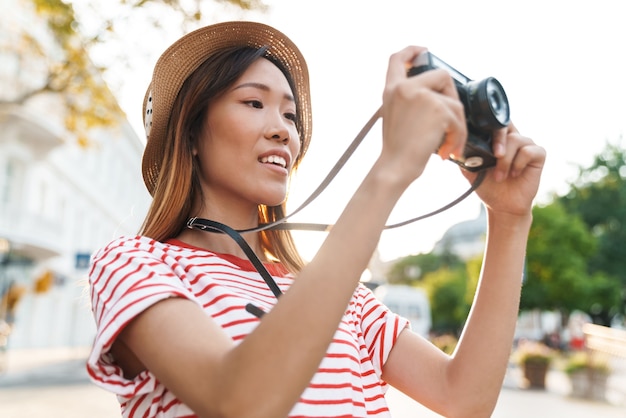 Portret van een tevreden Aziatische toeristenvrouw die op retro camera fotografeert terwijl ze op straat in de stad loopt