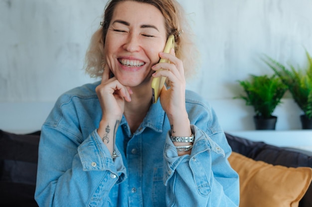 Portret van een tevreden blonde vrouw in een casual denim shirt die praat op een mobiele telefoon in een modern huis
