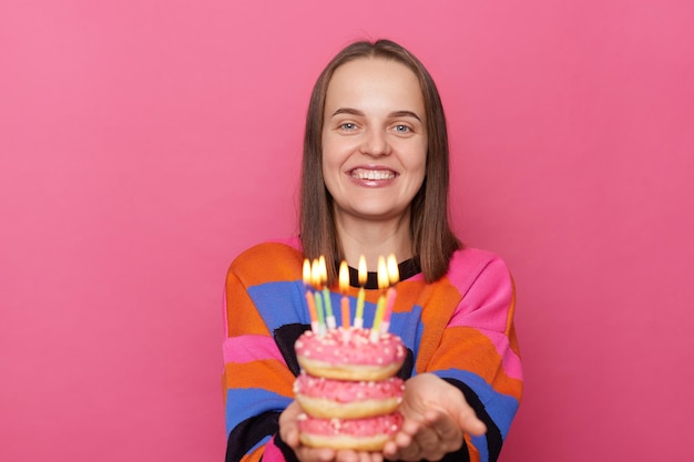 Portret van een tevreden bruinharige vrouw die heerlijke smakelijke donuts vasthoudt met brandende kaarsen met een trui die geïsoleerd staat over een roze achtergrond en naar de camera kijkt met een brede glimlach