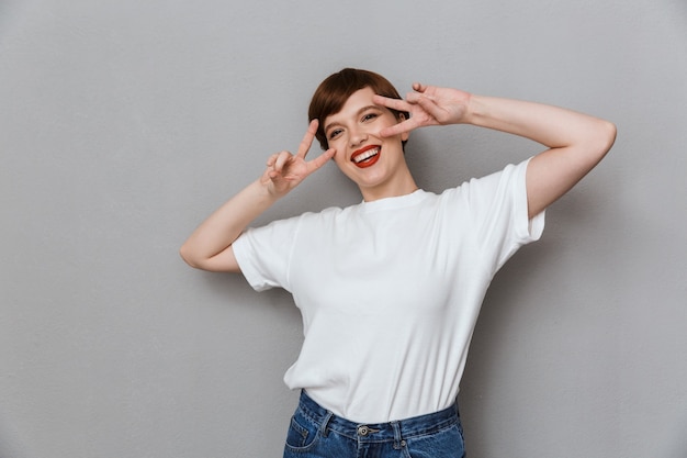 Portret van een tevreden brunette vrouw die een casual t-shirt draagt met een vredesteken met vingers geïsoleerd over een grijze muur