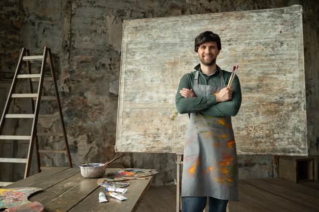 Portret van een tevreden jonge mannelijke kunstenaar die zijn wapens kruist met een borstel in zijn hand