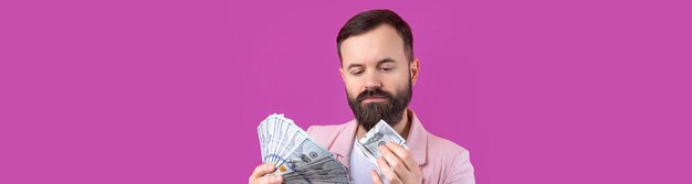Portret van een tevreden jonge zakenman met een baard gekleed in een roze jasje die ons dollarbankbiljetten toont tegen een rode studioachtergrond smaakgeur van geld