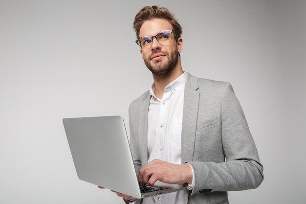 Portret van een tevreden knappe man in een bril die een laptop vasthoudt en gebruikt die over een witte muur wordt geïsoleerd