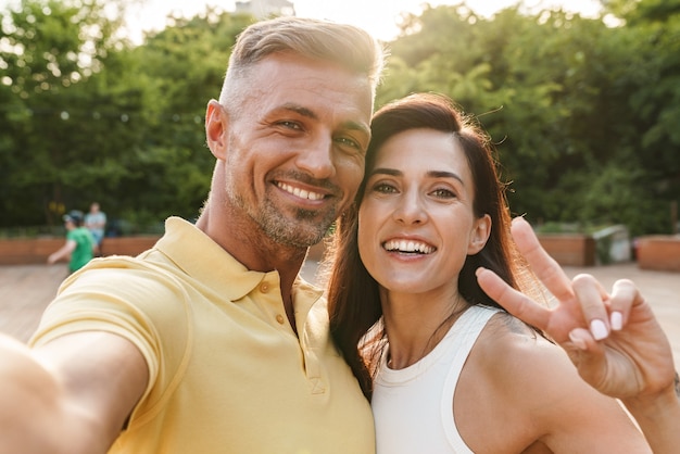 Portret van een tevreden man en vrouw van middelbare leeftijd die een selfie-foto maken en een vredesteken gebaren tijdens het wandelen in het zomerpark