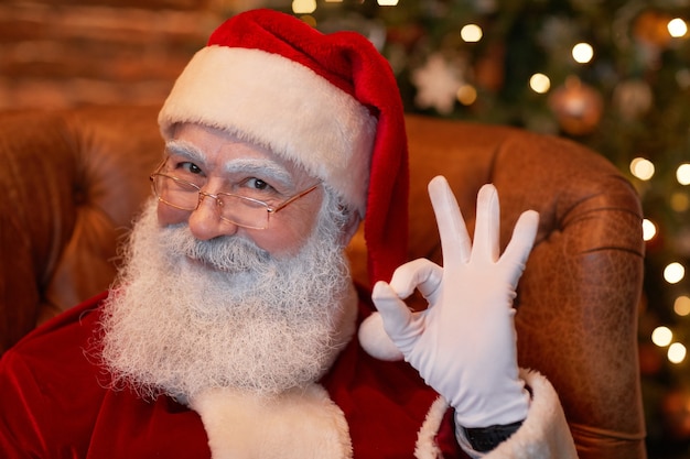 Portret van een tevreden oude kerstman met een witte baard in een bril die in een leunstoel zit en ok laat zien...