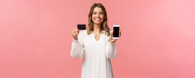 Portret van een tevreden schattig blond meisje in een witte jurk die je haar creditcard en mobiele telefoon laat zien