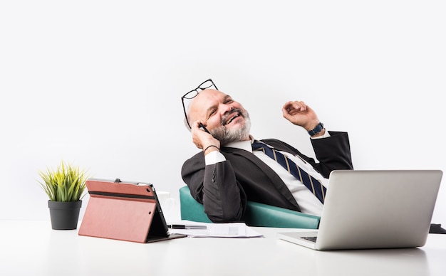 Portret van een tevreden senior Indiase Aziatische zakenman die op de werkplek achter de computer zit en naar de camera kijkt terwijl hij ontspant. Oude professionele man voeten op bureau