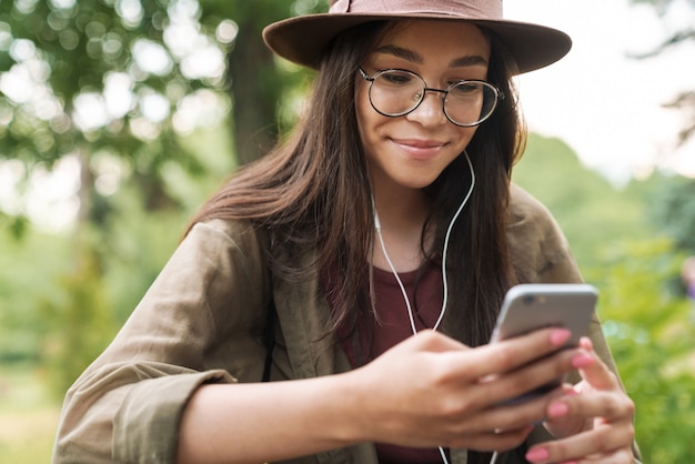 Portret van een tevreden vrouw met lang donker haar met een hoed en bril met oortelefoons en smartphone in groen park