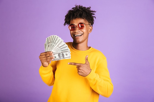 Portret van een tiener Afro-Amerikaanse man met een zonnebril die glimlacht terwijl hij contant geld dollars vasthoudt, geïsoleerd over violette achtergrond