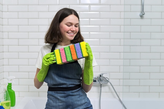 Portret van een tienermeisje dat in de badkamer schoonmaakt. Tiener in schorthandschoenen met sponzen. Schoonmaak, netheid thuis, service, jongerenconcept