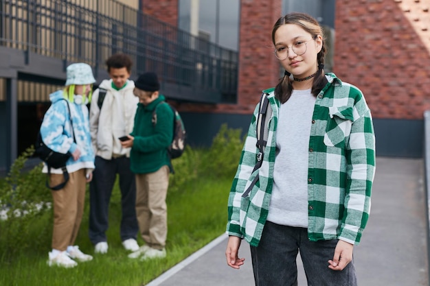 Foto portret van een tienermeisje met een bril die naar een camera kijkt die op het schoolplein staat met haar vrienden in