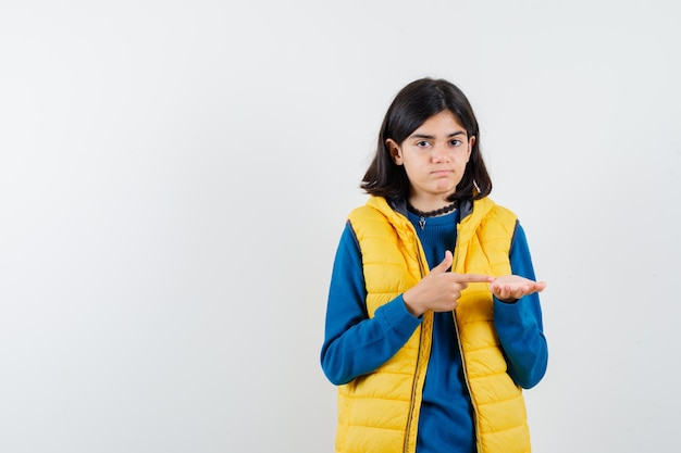 Portret van een tienermeisje met halflang haar in een blauwe trui en een geel vest dat denkt en een andere hand laat zien die op een witte achtergrond wordt geïsoleerd