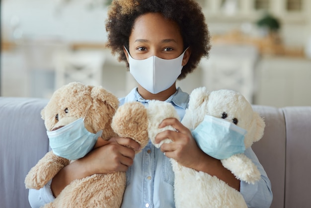 Portret van een tienermeisje van gemengd ras met een beschermend gezichtsmasker dat naar de camera kijkt en teddy vasthoudt