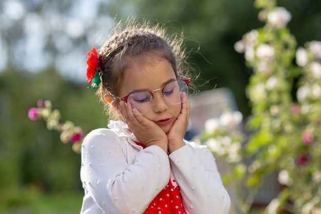 Portret van een triest mooi klein meisje in glazen, in een witte jas, haar wangen met haar handen vast te houden