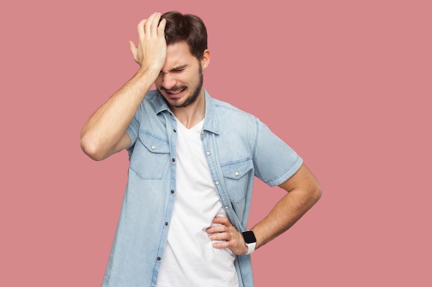 Portret van een trieste alleenstaande verliezer, bebaarde jongeman in een blauw casual stijlshirt dat zijn hoofd naar beneden houdt en nadenkt wat te doen. indoor studio opname, geïsoleerd op roze achtergrond.