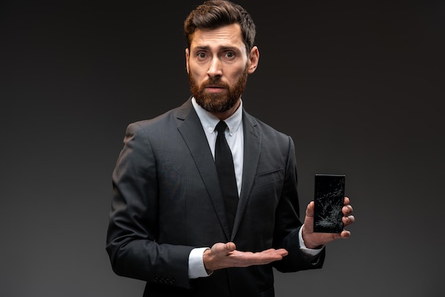 Portret van een trieste man in een formeel pak die een smartphone vasthoudt met een gebroken scherm dat op een zwarte achtergrond staat en naar de camera kijkt