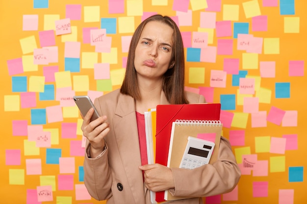 Portret van een trieste, overstuur, teleurgestelde vrouw in een beige jas die tegen een gele muur staat met kleurrijke memokaarten met papieren mappen en een rekenmachine die een mobiele telefoon gebruikt die slecht nieuws leest