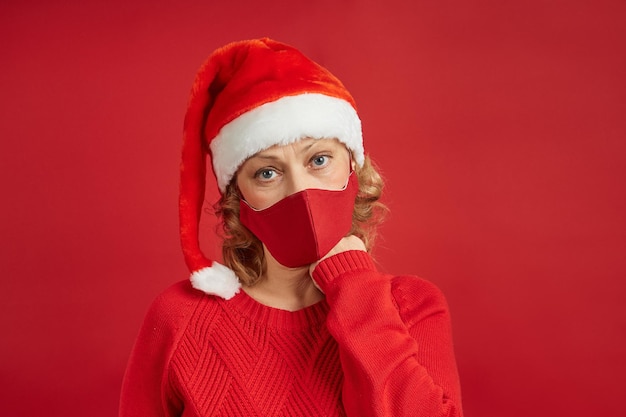 Portret van een trieste vrouw in een kerstmuts met een beschermend masker op een rode achtergrond.