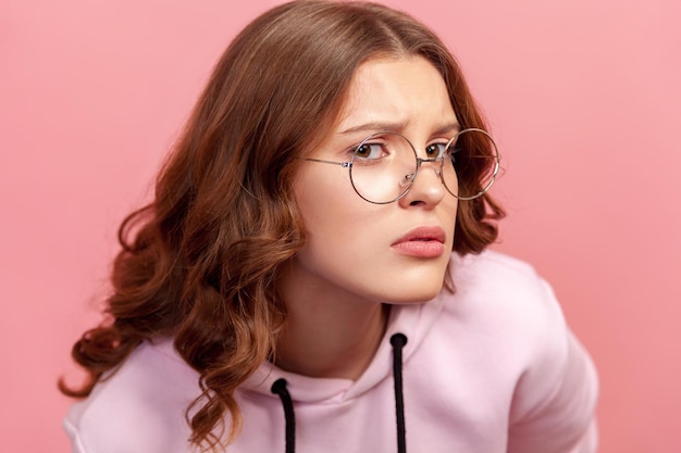 Portret van een twijfelende jonge vrouw met krullend haar in een bril die naar de camera kijkt met onzekere ogen en gefronste wenkbrauwen, onzeker over de keuze. Indoor studio-opname, geïsoleerd op roze achtergrond