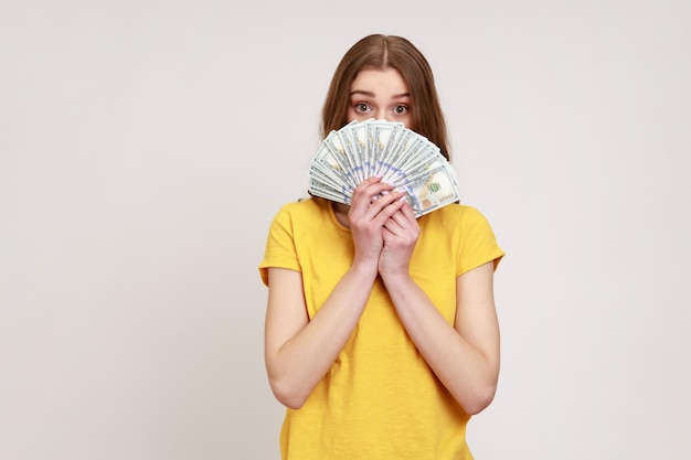 Portret van een verbaasd meisje in een casual geel T-shirt dat de helft van het gezicht bedekt met geld en uit de dollars gluurt, opgewonden om veel geld vast te houden. Indoor studio opname geïsoleerd op een grijze achtergrond.