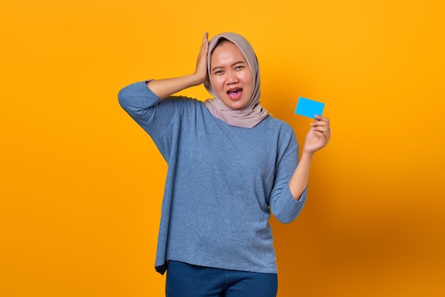 Portret van een verbaasde Aziatische vrouw die haar hand op haar hoofd houdt en creditcard toont over gele achtergrond