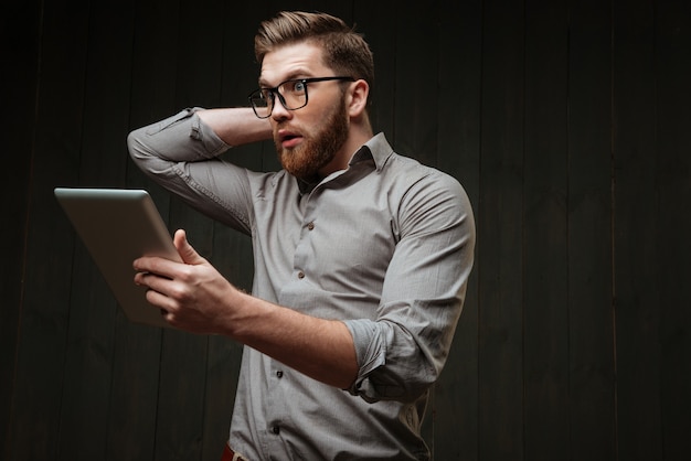 Portret van een verbaasde jonge man in een bril die een tabletcomputer vasthoudt en gebaren maakt geïsoleerd op een zwart houten oppervlak