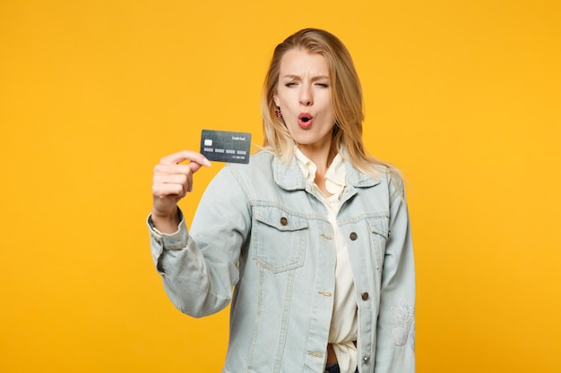 Portret van een verbaasde, verbaasde jonge vrouw in denim vrijetijdskleding met een creditcard geïsoleerd op een felgele oranje muurachtergrond in de studio. Mensen levensstijl concept. Bespotten kopie ruimte.