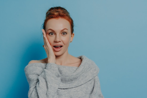 Foto portret van een verbaasde verbaasde roodharige vrouw die naar de camera kijkt met een geïmponeerde gezichtsuitdrukking, zich gelukkig voelt na het horen van onverwacht nieuws, geïsoleerd op een blauwe studioachtergrond, een vrouw die geschokt is