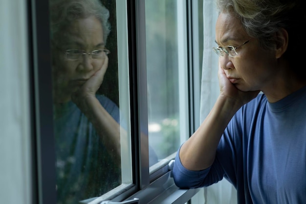 Portret van een verdrietige en bezorgde Aziatische oudere vrouw die thuis door het raam kijkt