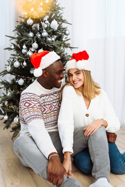 Portret van een verliefd stel in kerstmutsen die bij de boom zitten. Schittering van de gele slinger. Zwarte man knuffelt een blanke vrouw