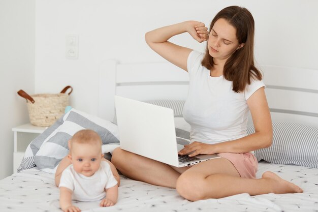 Portret van een vermoeide, uitgeputte vrouw die met haar dochtertje op bed zit en online werkt, laptop vasthoudt en handen uitrekt, lange uren online werkt.