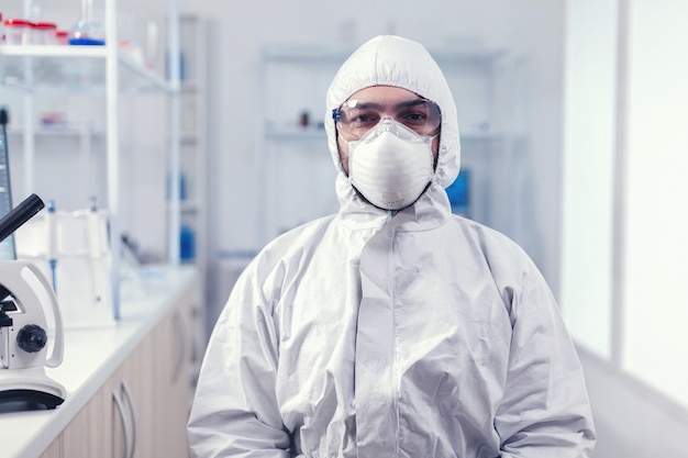 Portret van een vermoeide wetenschapper die een overall draagt met een veiligheidsbril die naar de camera kijkt in het medicijnlaboratorium. Overwerkte onderzoeker gekleed in beschermend pak tegen infectie met coronavirus tijdens