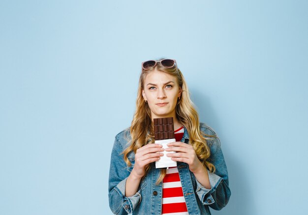 Portret van een verrast jong meisje dat over blauwe achtergrond wordt geïsoleerd, die chocolade houdt.