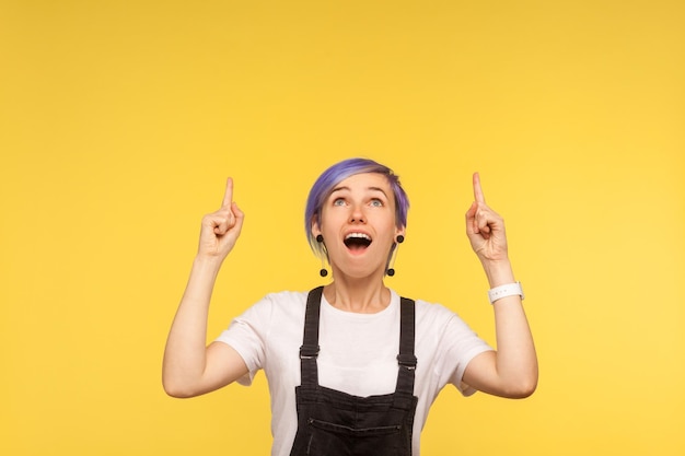 Portret van een verrast modieus meisje met violet kort haar in een denim overall die naar de vrije ruimte wijst voor reclame, en er geschokt uitziet. geïsoleerd op gele achtergrond, studio-opname, kopieer ruimte