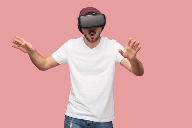 Portret van een verraste, bebaarde jonge hipster-man in een wit overhemd en een casual hoed die staat, vr draagt en video kijkt op de simulator met opgeheven armen. binnen, geïsoleerd, studio-opname, roze achtergrond