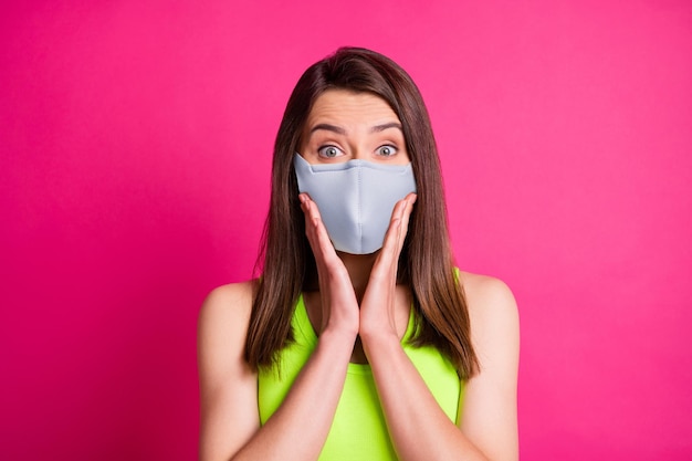 Portret van een verraste brunette dame draagt een blauw stoffen gezichtsmasker dat op een felroze achtergrond wordt geïsoleerd