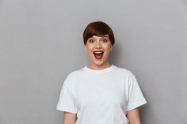 Portret van een verraste brunette vrouw die een casual t-shirt draagt dat verwondering uitdrukt en lacht aan de voorkant, geïsoleerd over een grijze muur