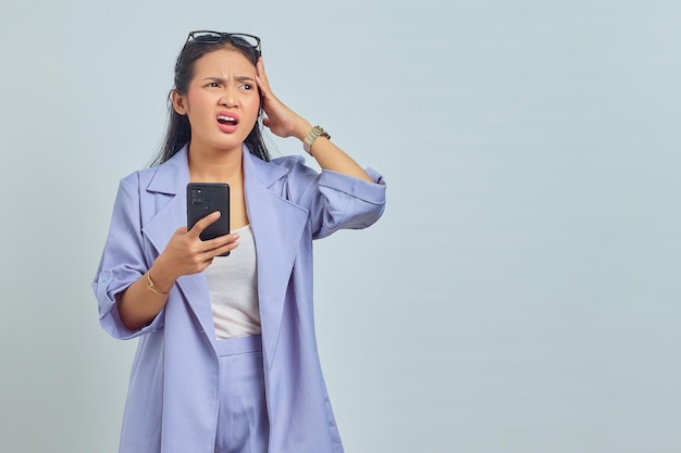 Portret van een verraste jonge Aziatische vrouw die een mobiele telefoon gebruikt met een handgebaar dat het hoofd vasthoudt en wegkijkt op een witte achtergrond