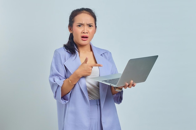 Portret van een verraste jonge Aziatische vrouw die met de vingers naar een laptop wijst en naar een camera kijkt die op een witte achtergrond is geïsoleerd