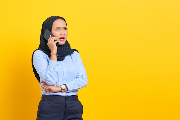 Portret van een verraste jonge Aziatische vrouw die op een mobiele telefoon praat en wegkijkt geïsoleerd op een gele achtergrond