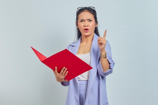 Portret van een verraste jonge Aziatische zakenvrouw in een pak die een documentmap vasthoudt en een vinger naar boven wijst, geïsoleerd op een paarse achtergrond