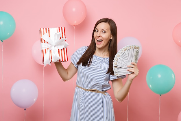 Portret van een verraste jonge vrouw in blauwe jurk met bundel veel dollars contant geld en rode doos met cadeau aanwezig op roze achtergrond met kleurrijke luchtballonnen. Verjaardagsfeestje concept.