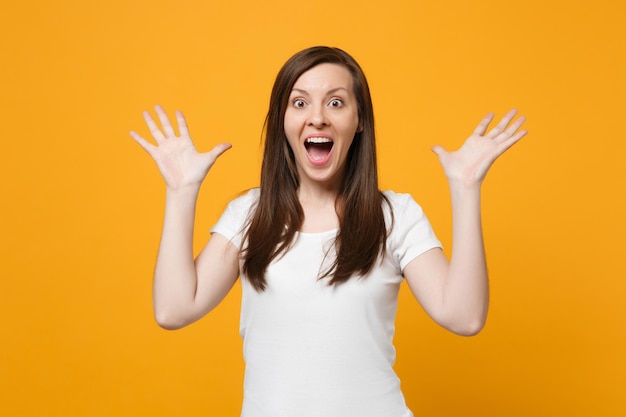 Portret van een verraste jonge vrouw in witte vrijetijdskleding die de mond openhoudt, handen spreidt geïsoleerd op een felgele oranje muurachtergrond in de studio. Mensen levensstijl concept. Bespotten kopie ruimte.