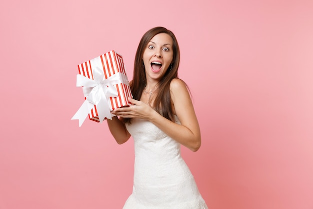 Portret van een verraste opgewonden vrouw in een kanten witte jurk met een rode doos met cadeau, aanwezig